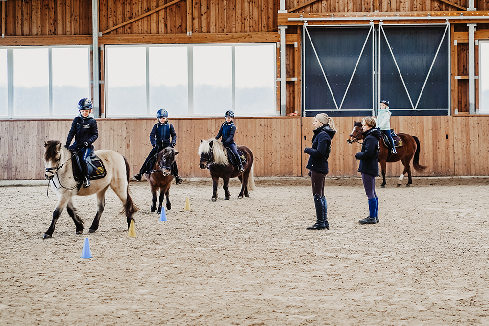 Kleine Gruppe von Kindern auf Ponnys in Reithalle, Trainerinnen beobachten
