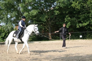Reitschülerin auf weißem Pferd bein Longenunterricht, Mitarbeiterin hält Leine von Pferd