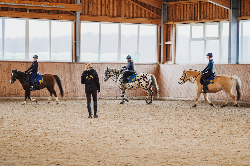 Kleine Gruppe von Teitschülerinnnen auf Pferden in Reithalle, Trainerin beobachtet