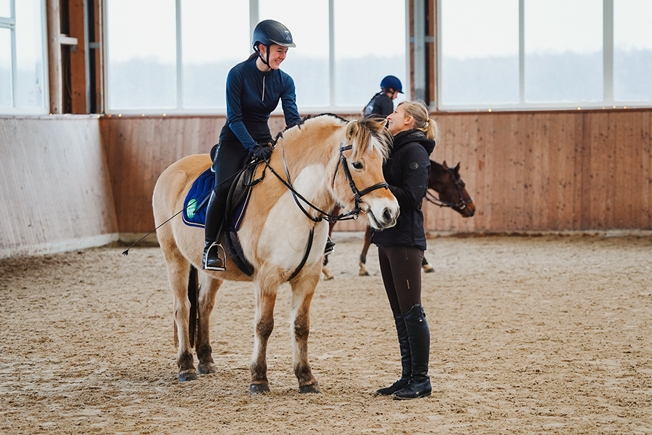In Reithalle: Reitschülerin auf Pferd spricht lächelnd mit Trainerin