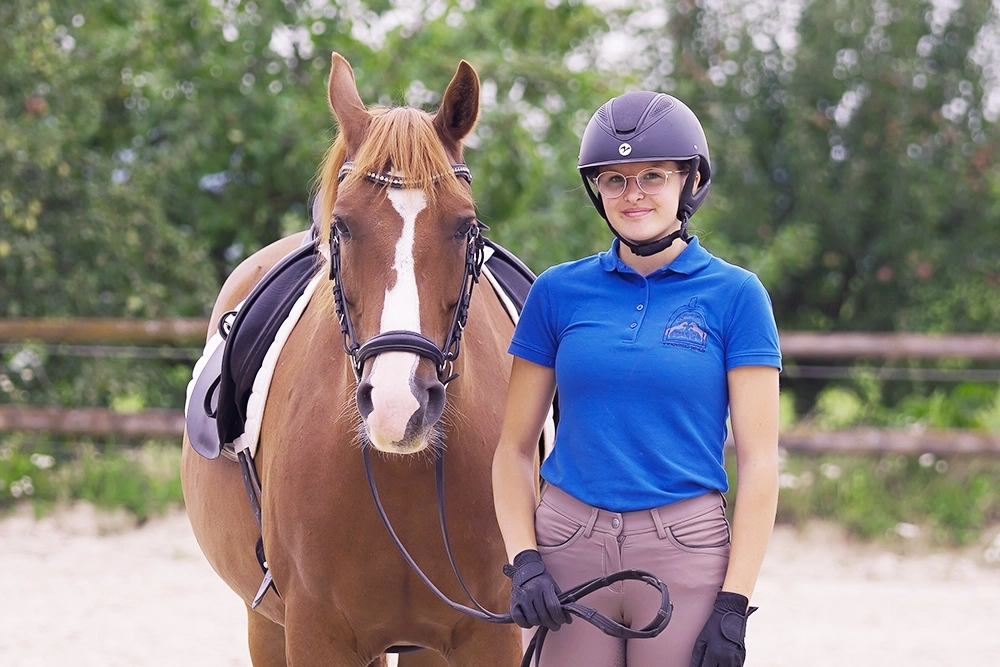 Lea steht neben braunem Pferd, hält Zügel in Hand, trägt Reithelm